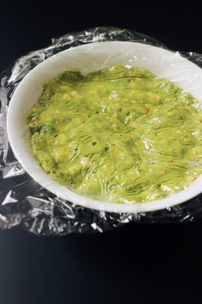 bowl of guacamole with plastic wrap on surface