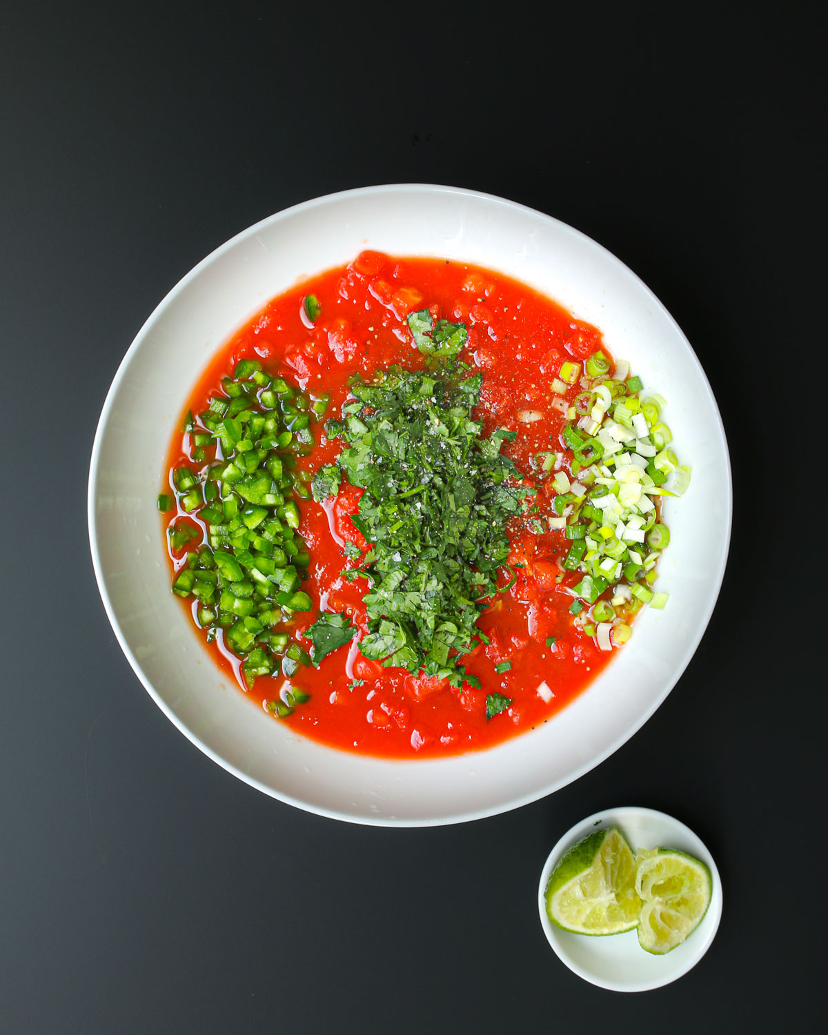tomatoes in a large white bowl with the chopped onion, cilantro, and jalapeno in lines atop, the lime wedges are in a small white dish nearby on the black table.