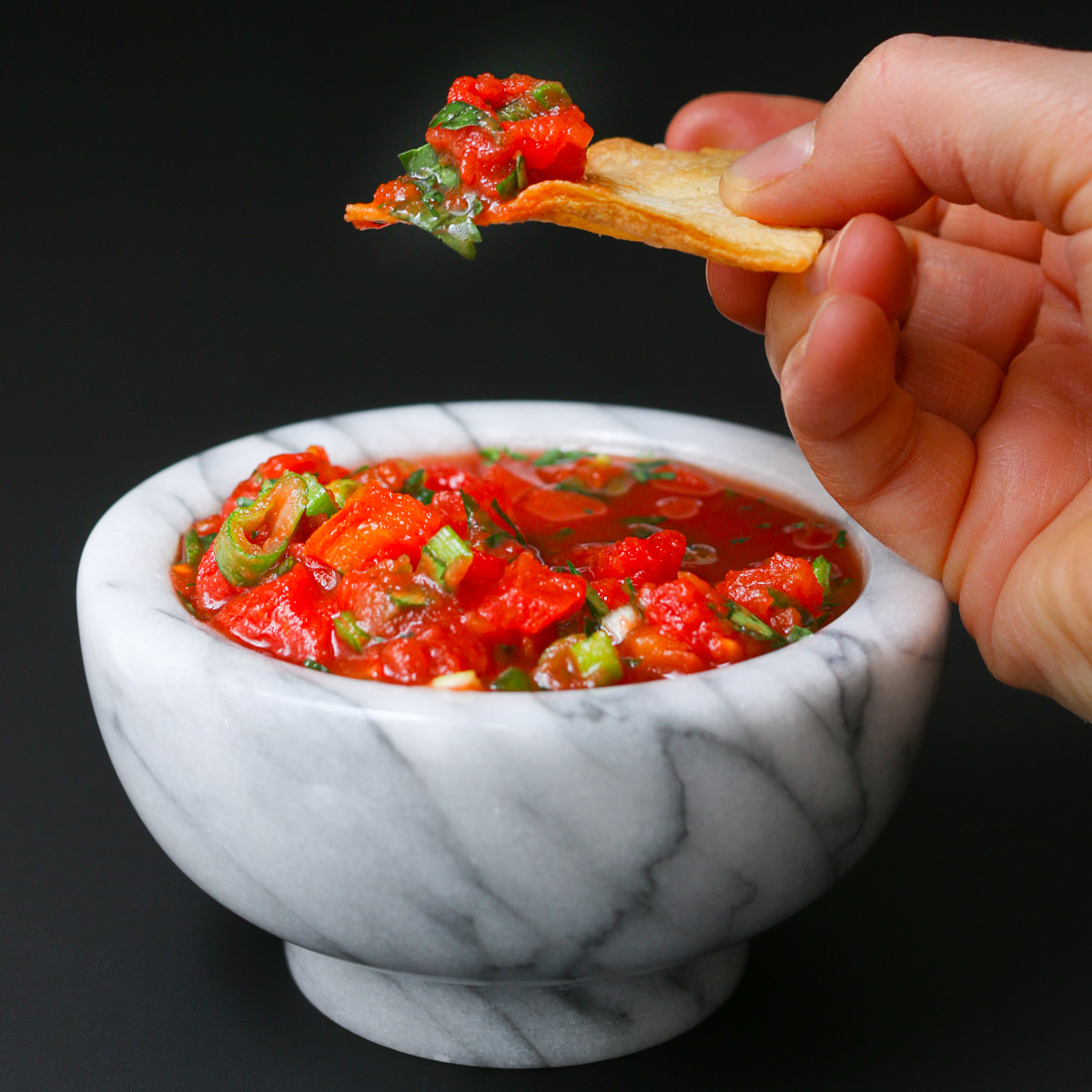 hand holding chip piled with salsa above a marble pestle of salsa.