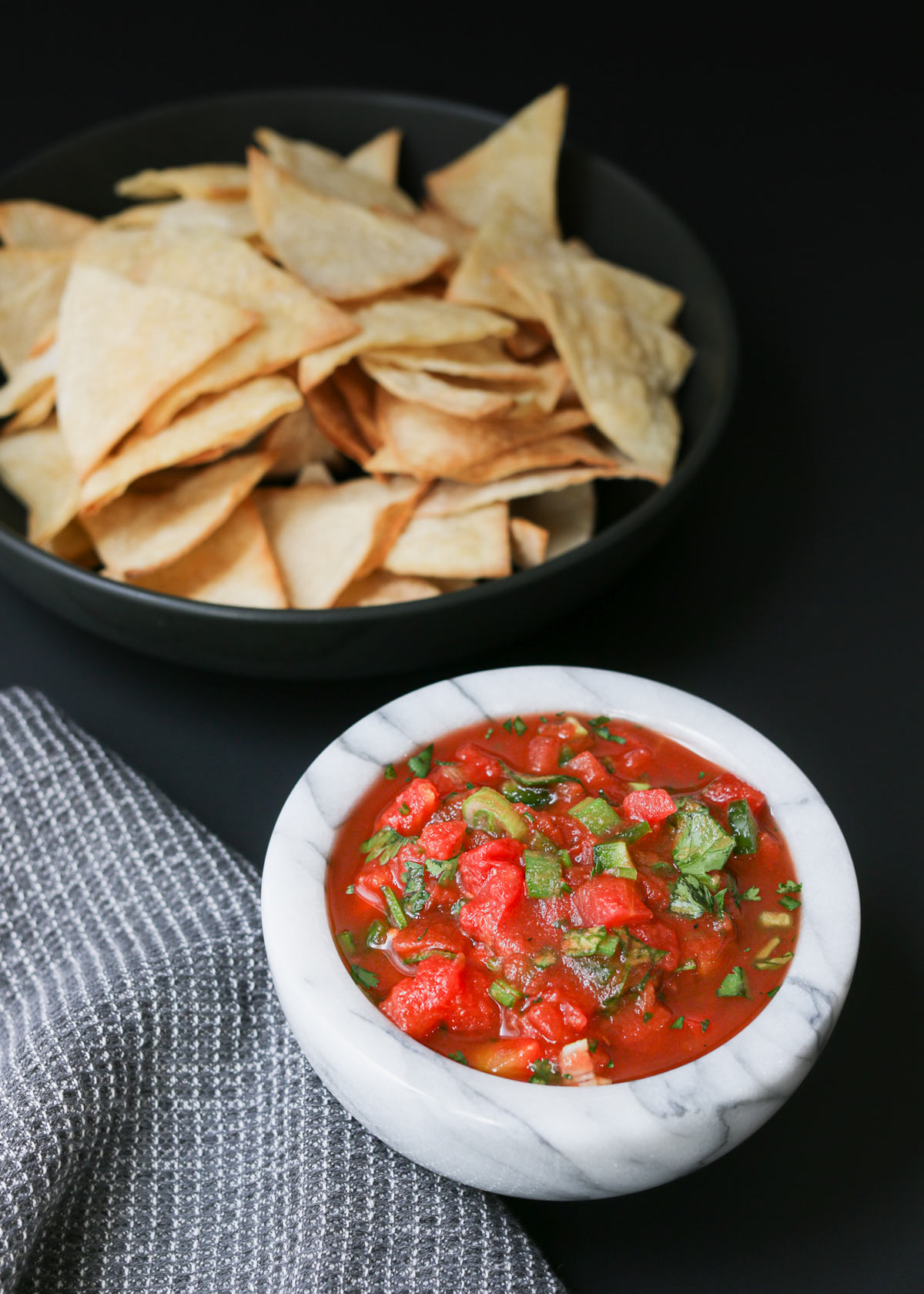 salsa in a marble bowl on a black table next to a gray cloth and a black bowl of tortilla chips.