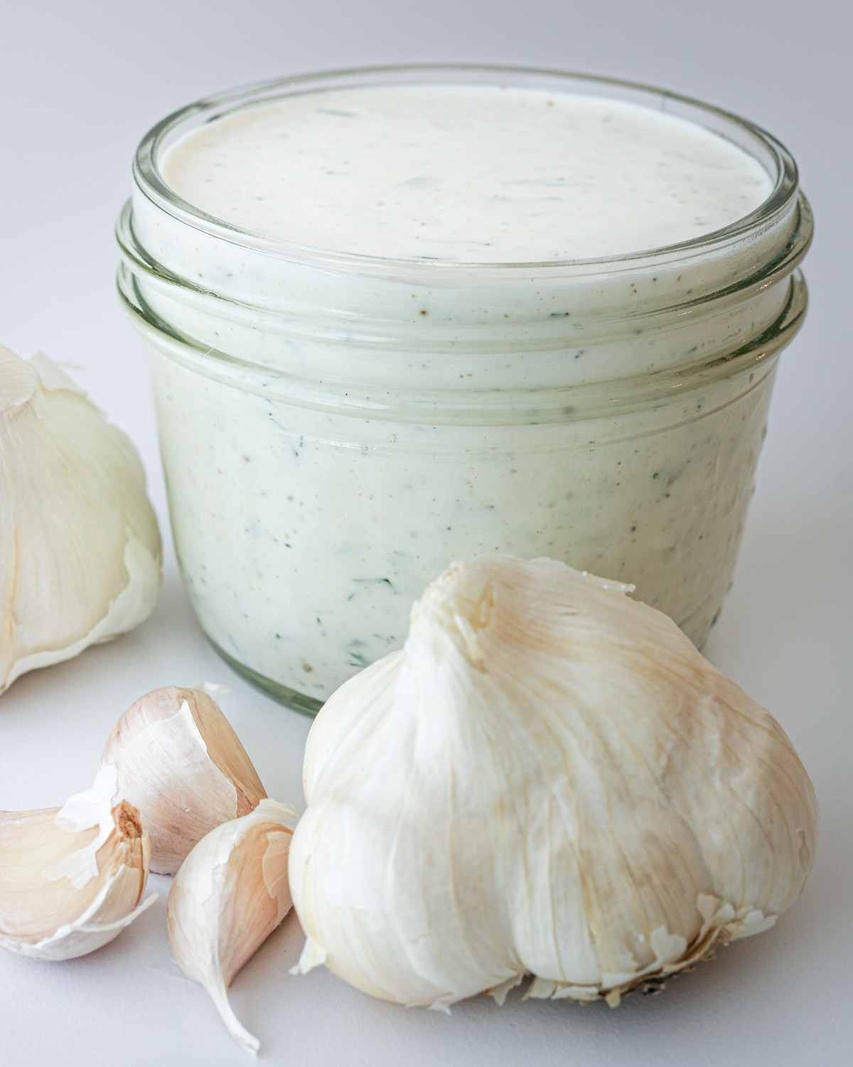 jar of creamy garlic dressing with cloves and heads of garlic in the foreground.