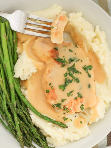 a bite of chicken on a fork over a plate of chicken and gravy with asparagus on the side.