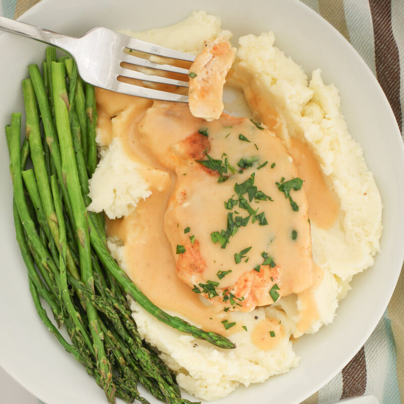 a bite of chicken on a fork over a plate of chicken and gravy with asparagus on the side.