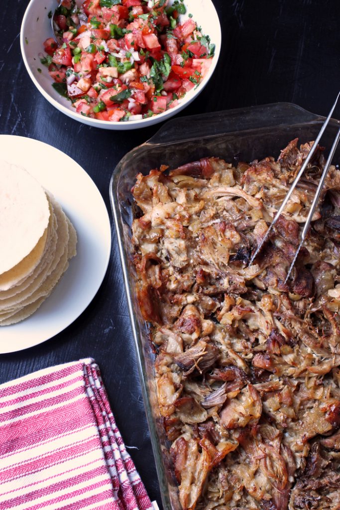 carnitas in glass dish next to stack of tortillas and bowl of salsa