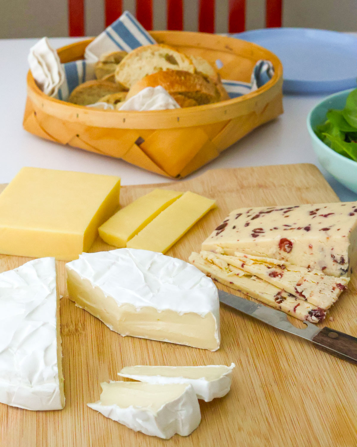 cheese board on table with bread basket and salad bowl nearby.