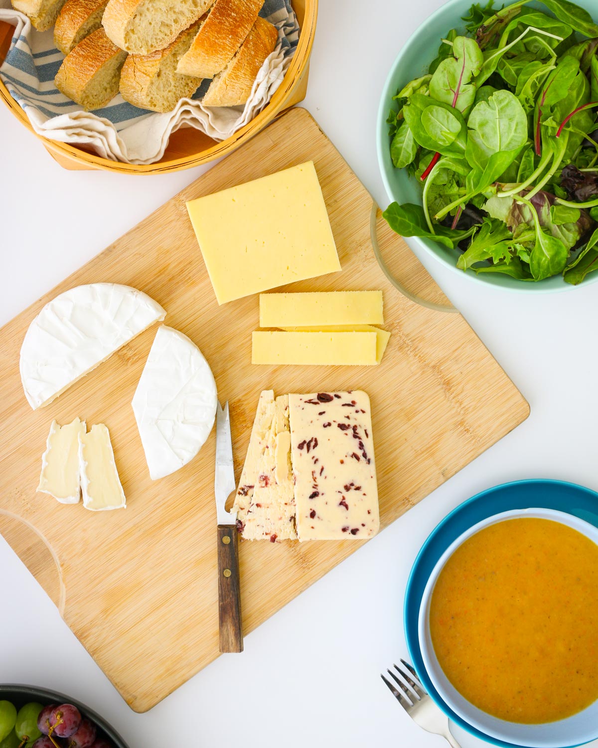 dinner table with a cheese plate in the center, surrounded by bread basket, soup bowl, salad bowl, and fruit.