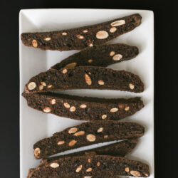 chocolate biscotti with almonds on a white platter on a black table.