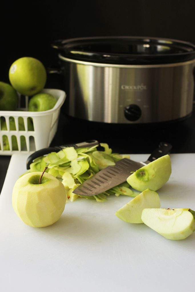 slowcooker next to basket of apples and cutting board with knife, peelings, and cut apples