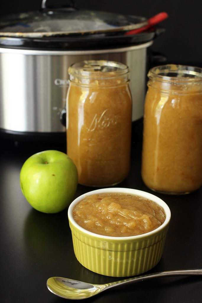 A bowl of applesauce on a table, next to jars and apple