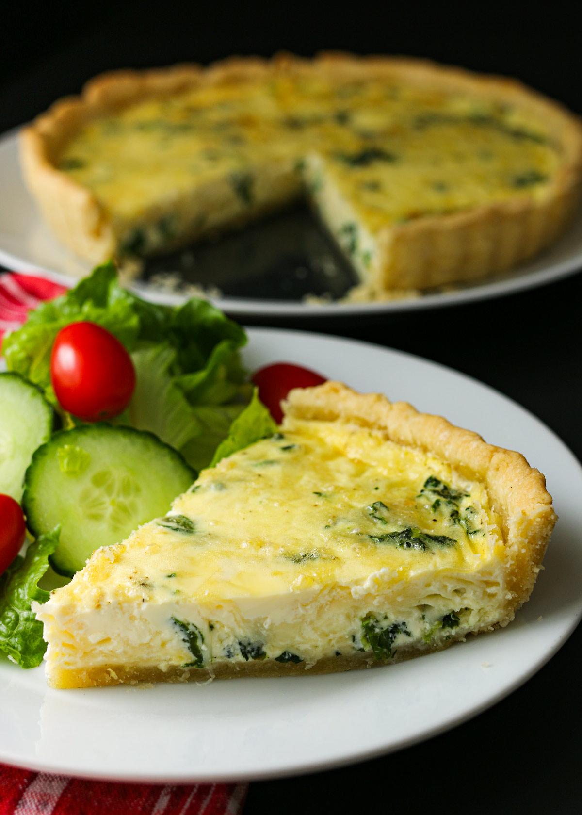 wedge of quiche on a plate with salad with larger quiche in background.