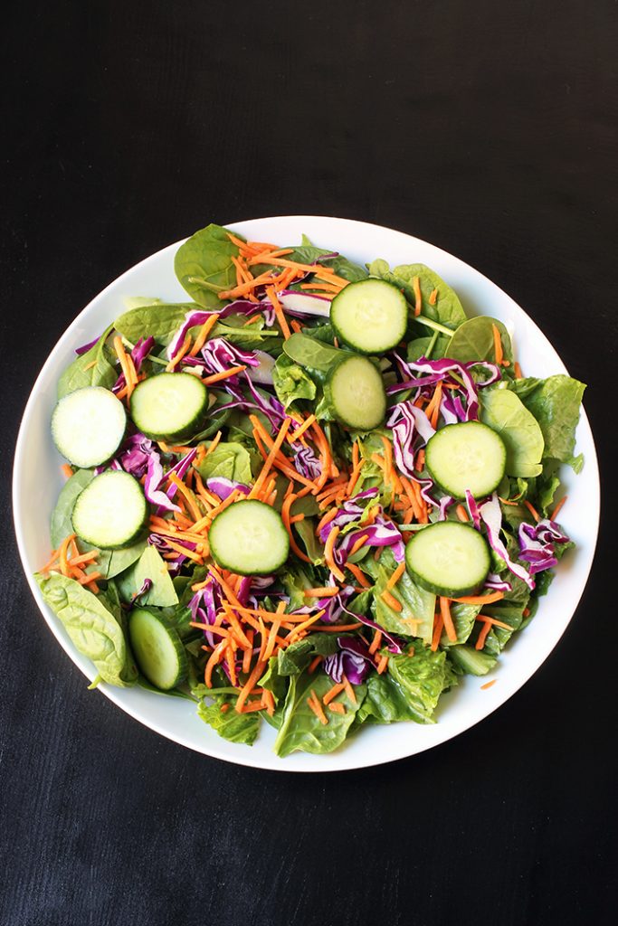 cucumbers and shredded carrots added to greens in salad bowl.