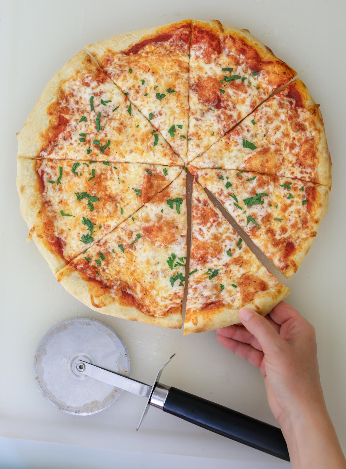 hand grabbing a slice of cheese pizza out of the whole pie, with a pizza cutter nearby.