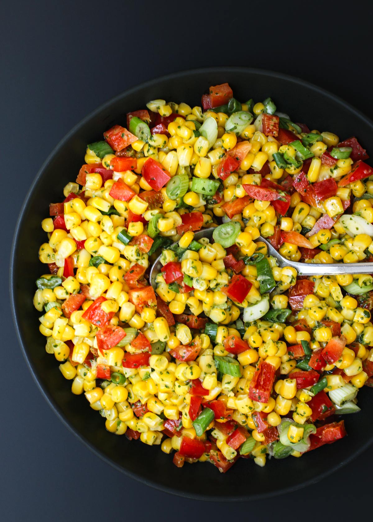 a full serving spoon resting in the center of a black bowl of corn salad.
