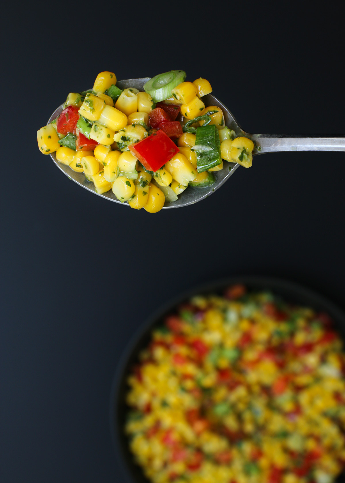 overhead shot of a spoonful of corn salad held above a black serving bowl on a black table.