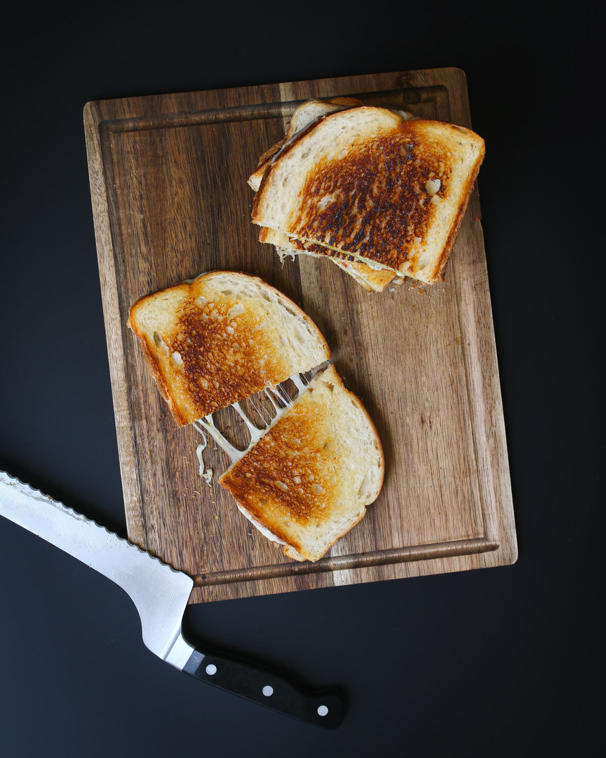 overhead shot of grilled sandwich cut in half with cheese pull and a knife and stacked sandwich on board nearby on black table.