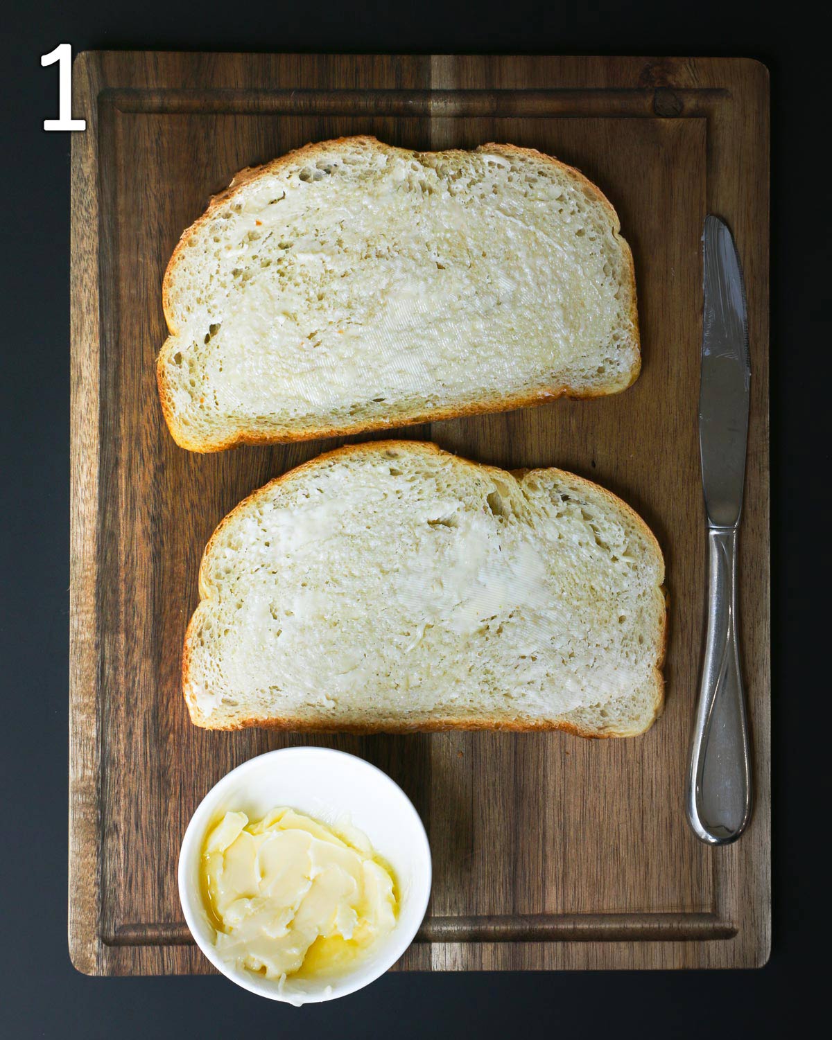 two slices of buttered bread on board with knife and dish of butter.