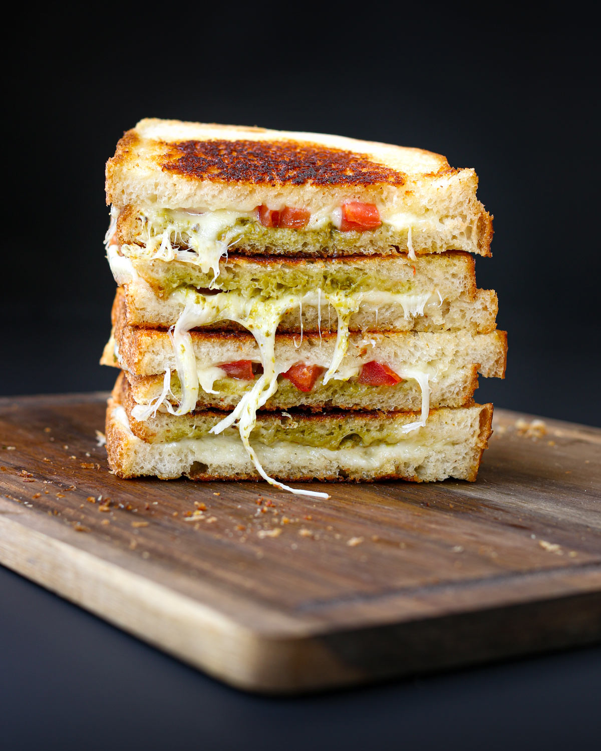 two pesto grilled cheese sandwiches cut and stacked on a wooden board with a black background.