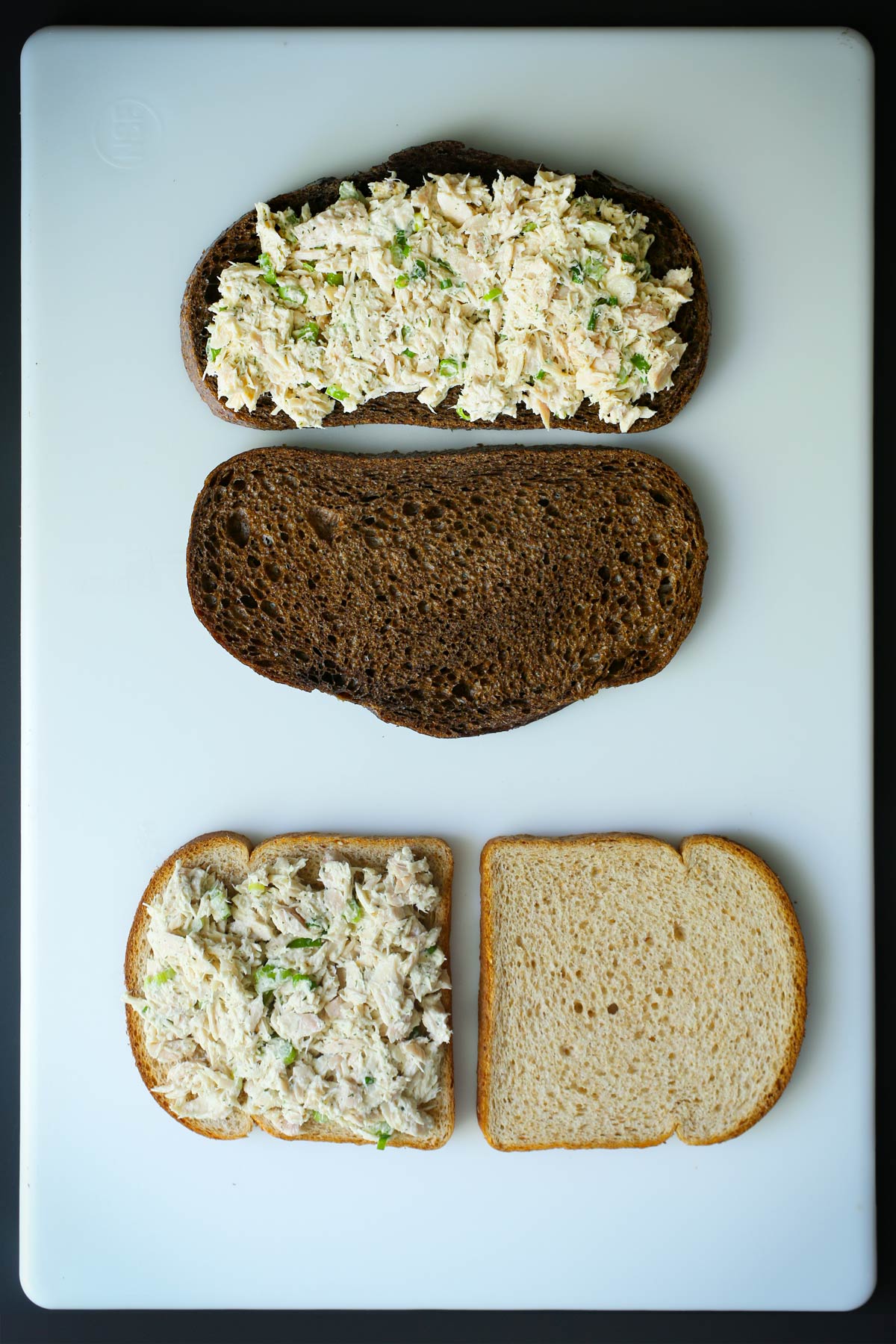 assembling tuna sandwiches on a white cutting board, using two kinds of sandwich bread.