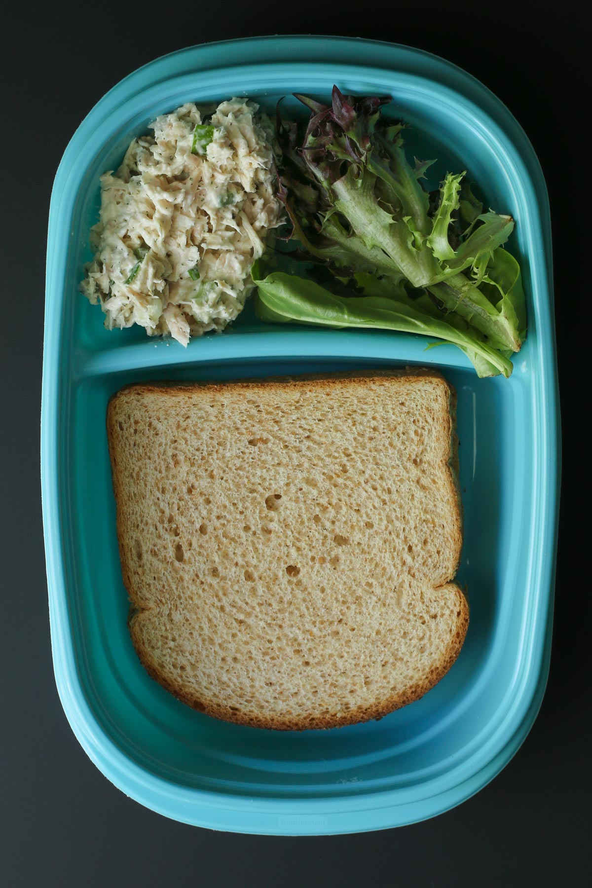 teal meal prep box with bread slices in one compartment and tuna salad and lettuce leaves in another.