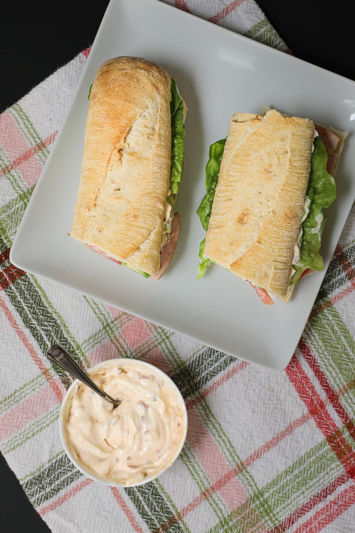 baguette sandwiches on a square white plate with a bowl of chipotle mayo nearby, all on a green and pink plaid cloth on a black table.