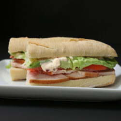 in front of a black backdrop are two portions of baguette sandwich with the chipotle mayo evident in a blob on the side.