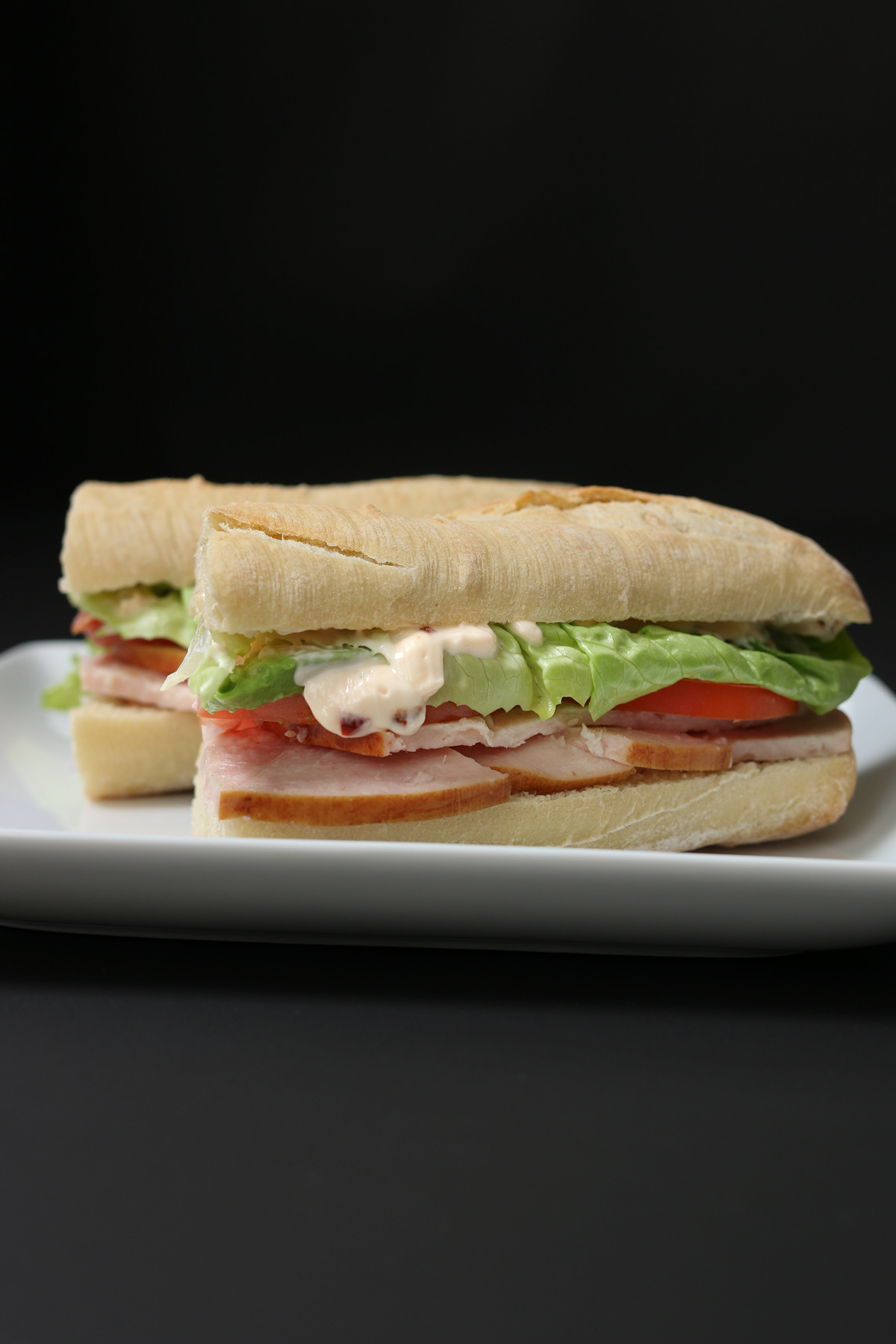two portions of turkey baguette sandwich on white square plate with black background.