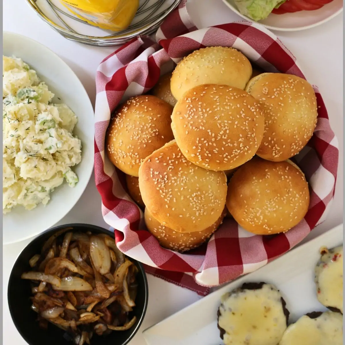a basket of hamburger buns next to a platter of burgers and toppings.