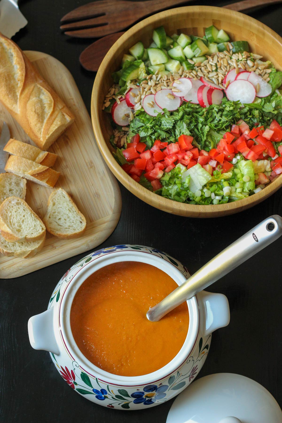 overhead shot of vegetable soup in tureen with ladle near a board of bread and a large salad.