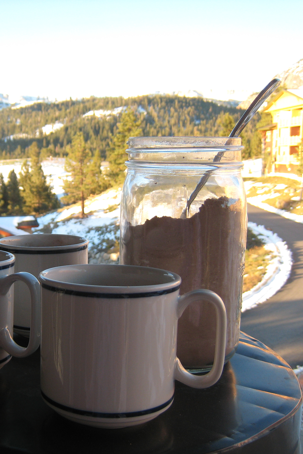 mugs and jar of cocoa mix on balcony pillar with snowy mountains in background