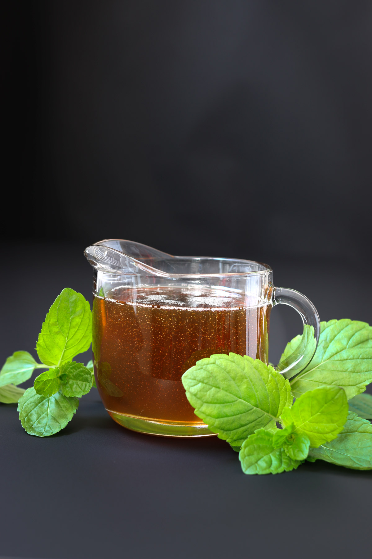 sideview of peppermint syrup in glass pitcher with sprigs of fresh mint nearby on black table.