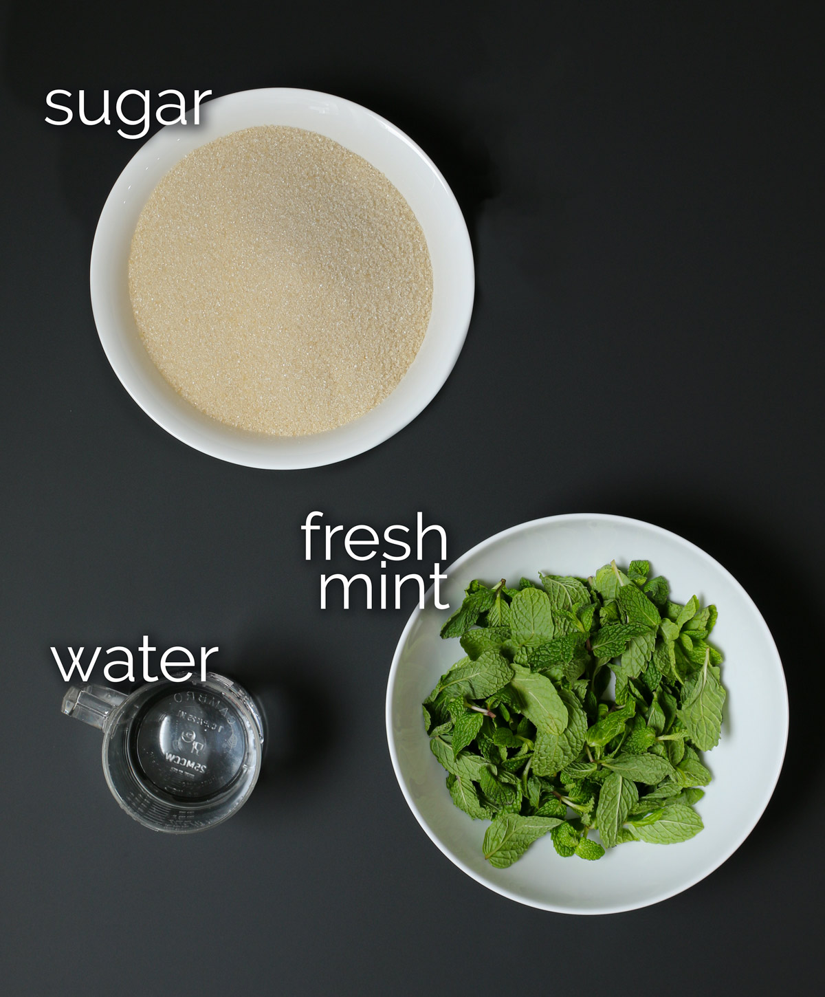 bowl of sugar next to clear cup of water and bowl of mint leaves on a black table.