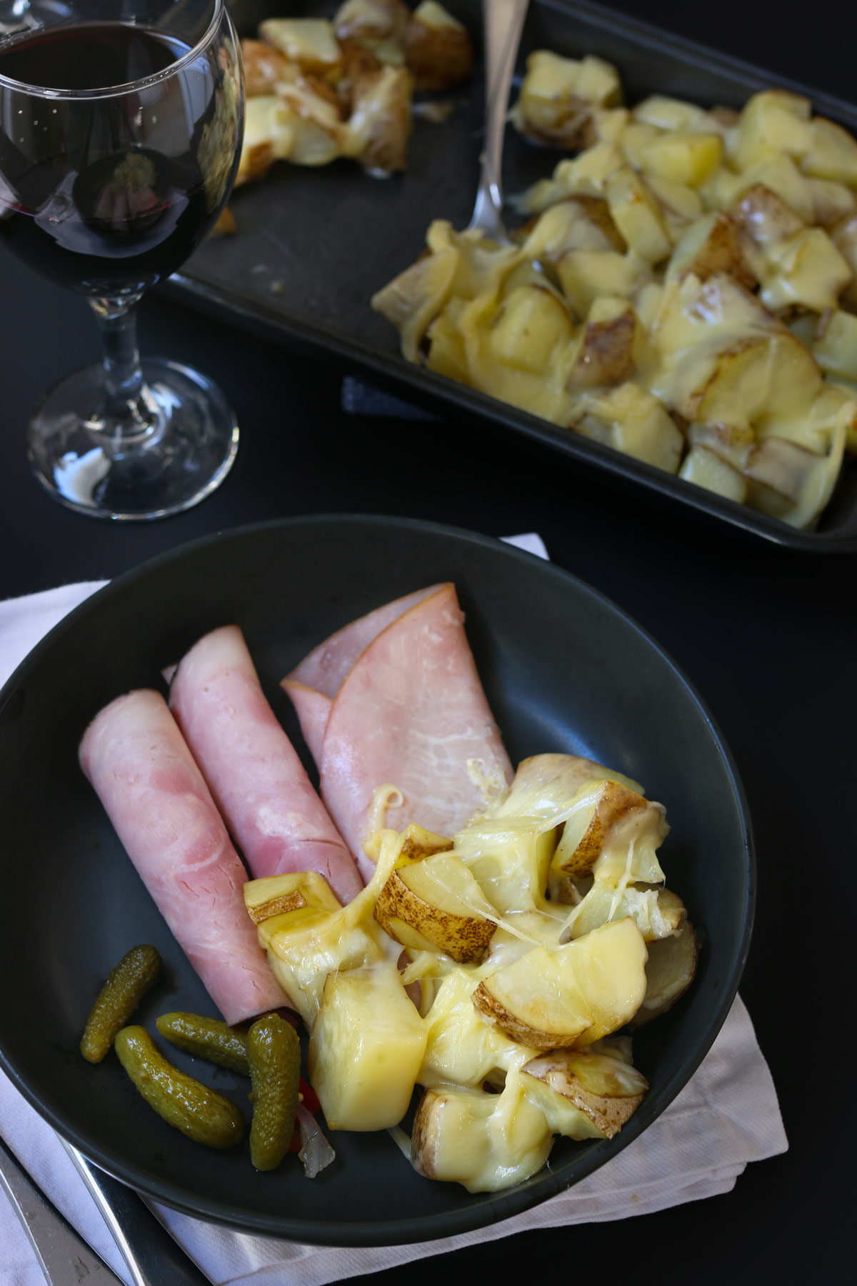 ham, raclette, potatoes, and pickles in black dish on a white napkin next to a glass of wine and the platter of raclette.