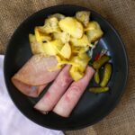 overhead shot of raclette melted over potatoes in a black dish with pickles and ham.