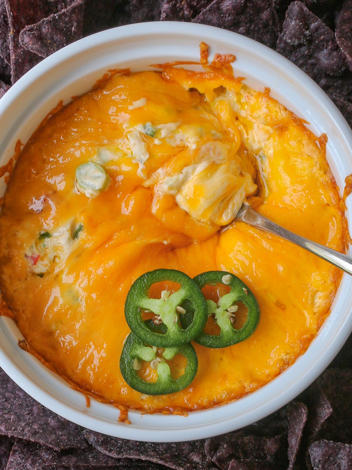 bowl of jalapeno cheese dip, with spoon swirled in the melted cheese, surrounded by blue corn tortilla chips.