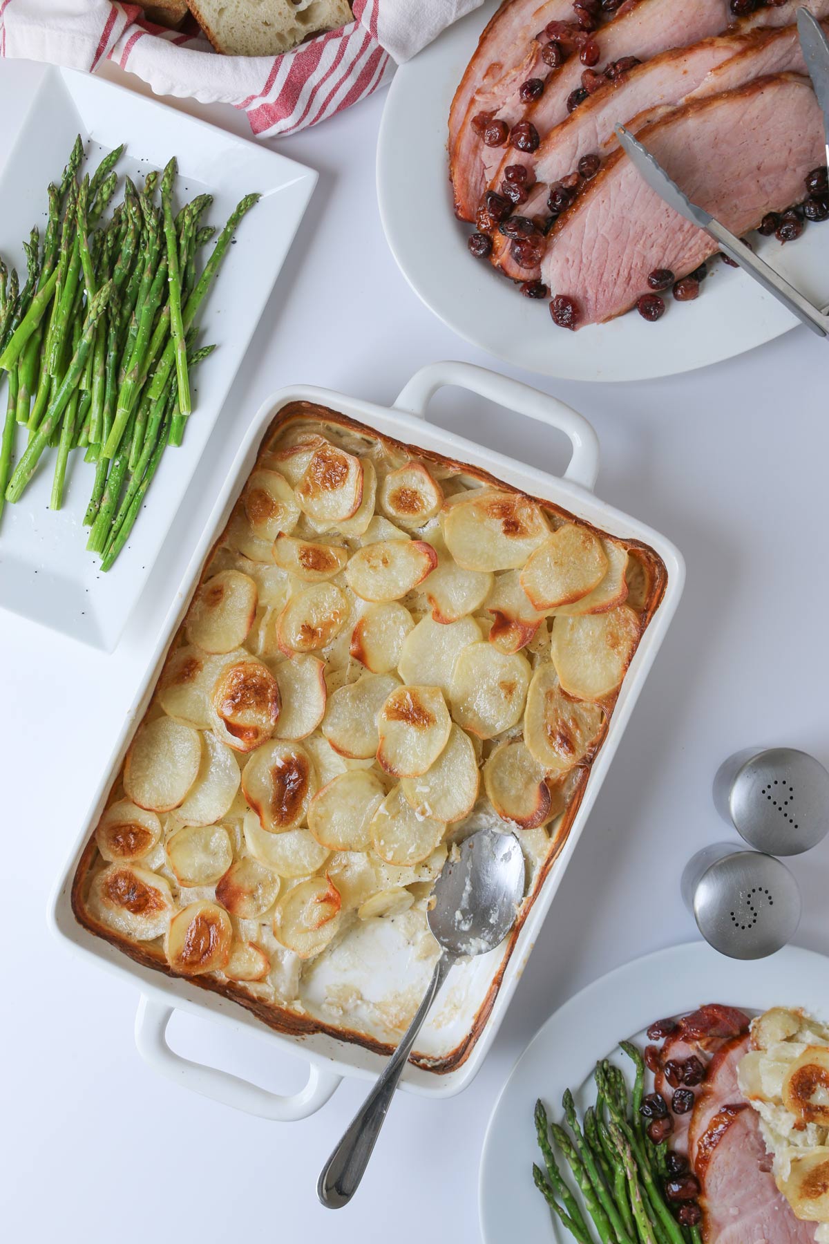 a dish of scalloped potatoes with a serving spoon on a table surrounded by other meal elements. 