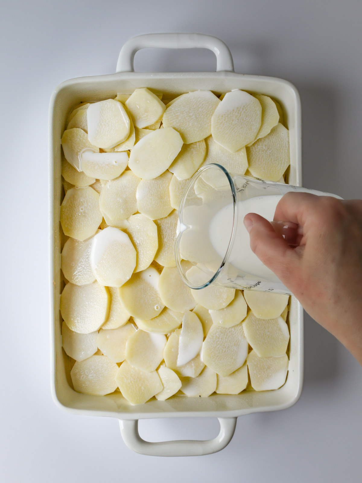 pouring half and half over layered potatoes in a baking dish.