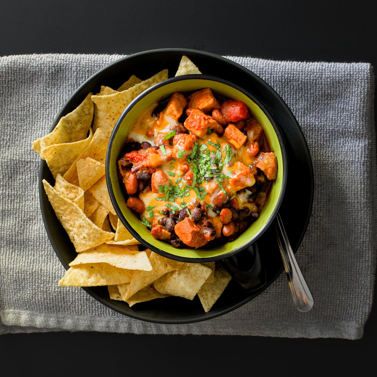 bowl of chili atop plate of chips, on a gray cloth and black table.