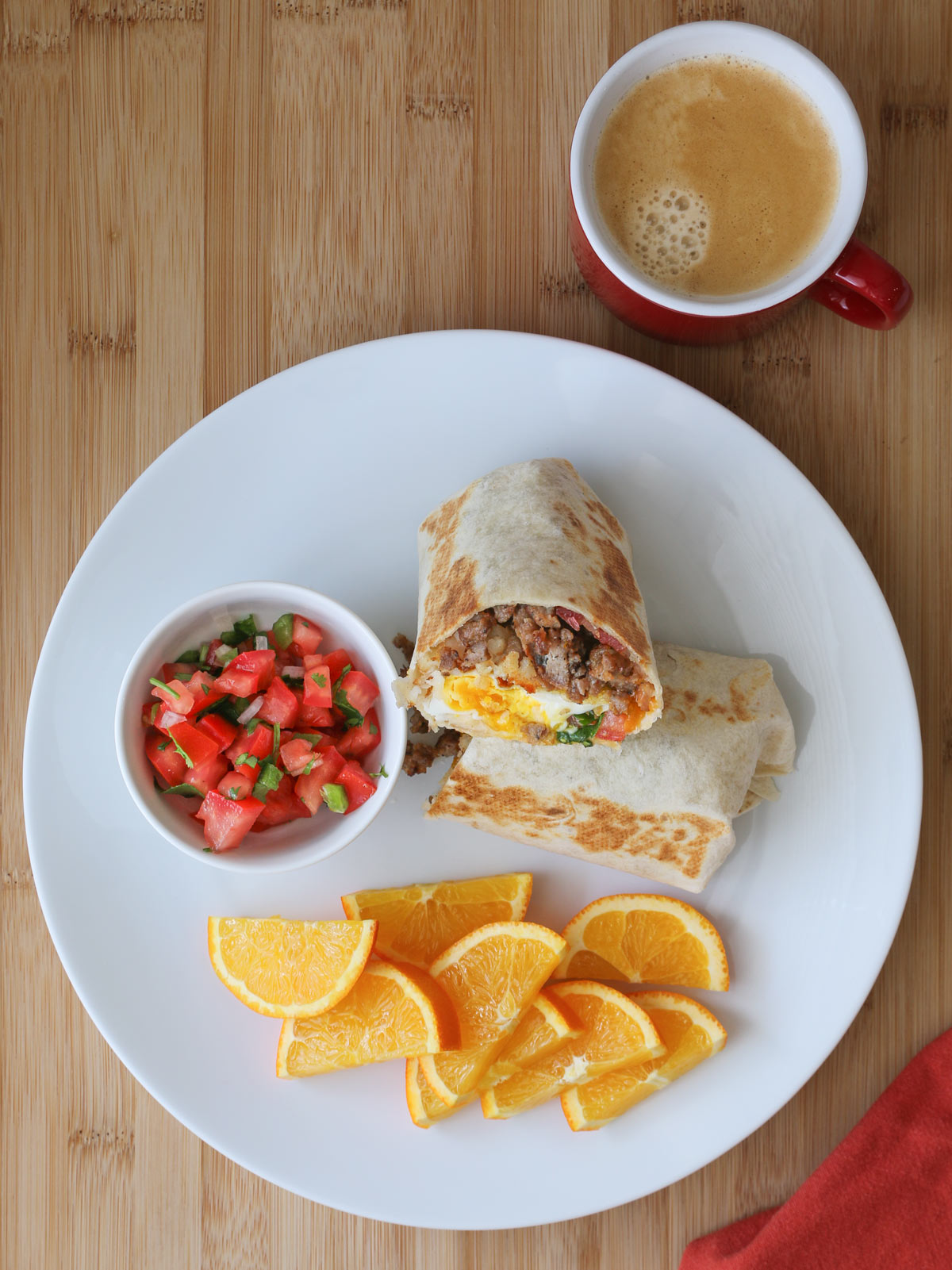 breakfast plate with burrito cut in half with orange slices and bowl of pico, cup of coffee nearby.