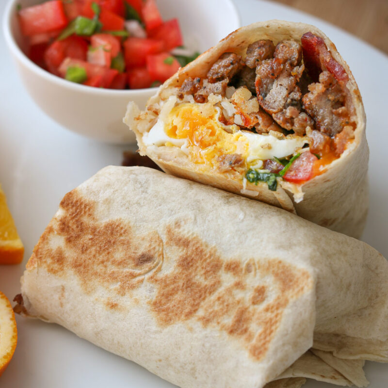 one half of a breakfast burrito leaning on the other half with cup of pico nearby.