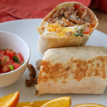 cut half of breakfast burrito leaning on second half on plate with pico and orange slices.