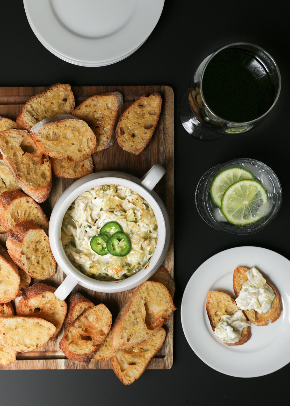 overhead shot of appetizer table with artichoke dip in tureen on a board of baguette toast, next to two party beverages and a plate with toast and dip.