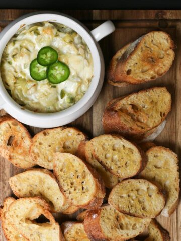 overhead shot of jalapeno artichoke dip in white tureen on brown wood board, surrounded with toasts for dipping.