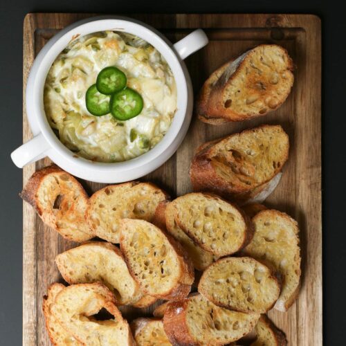 overhead shot of jalapeno artichoke dip in white tureen on brown wood board, surrounded with toasts for dipping.