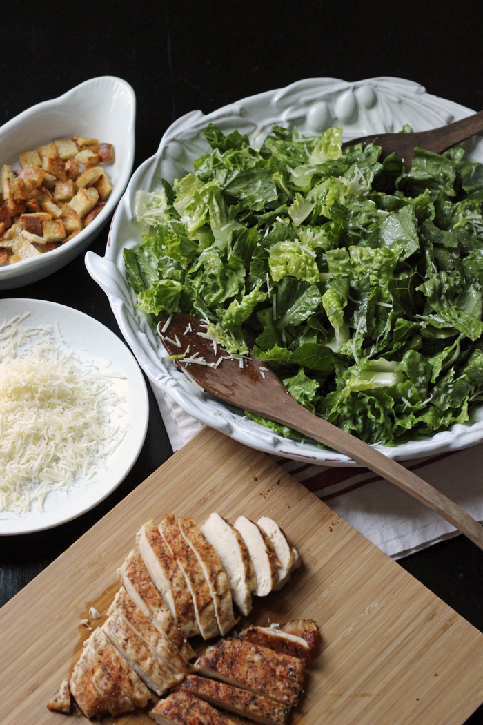 A bowl of romaine, bowl of croutons, cutting board of chicken ,and croutons