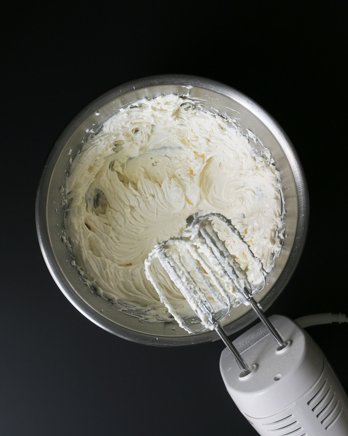 whipped cream cheese in metal bowl next to beaters of mixer.