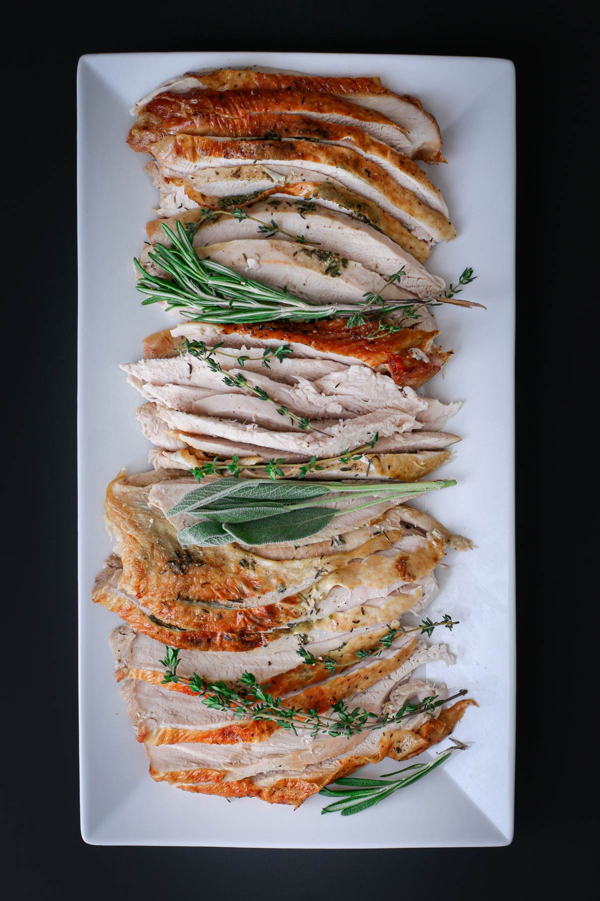 overhead shot of rectangle platter with sliced roast turkey breast and fresh herb sprigs.