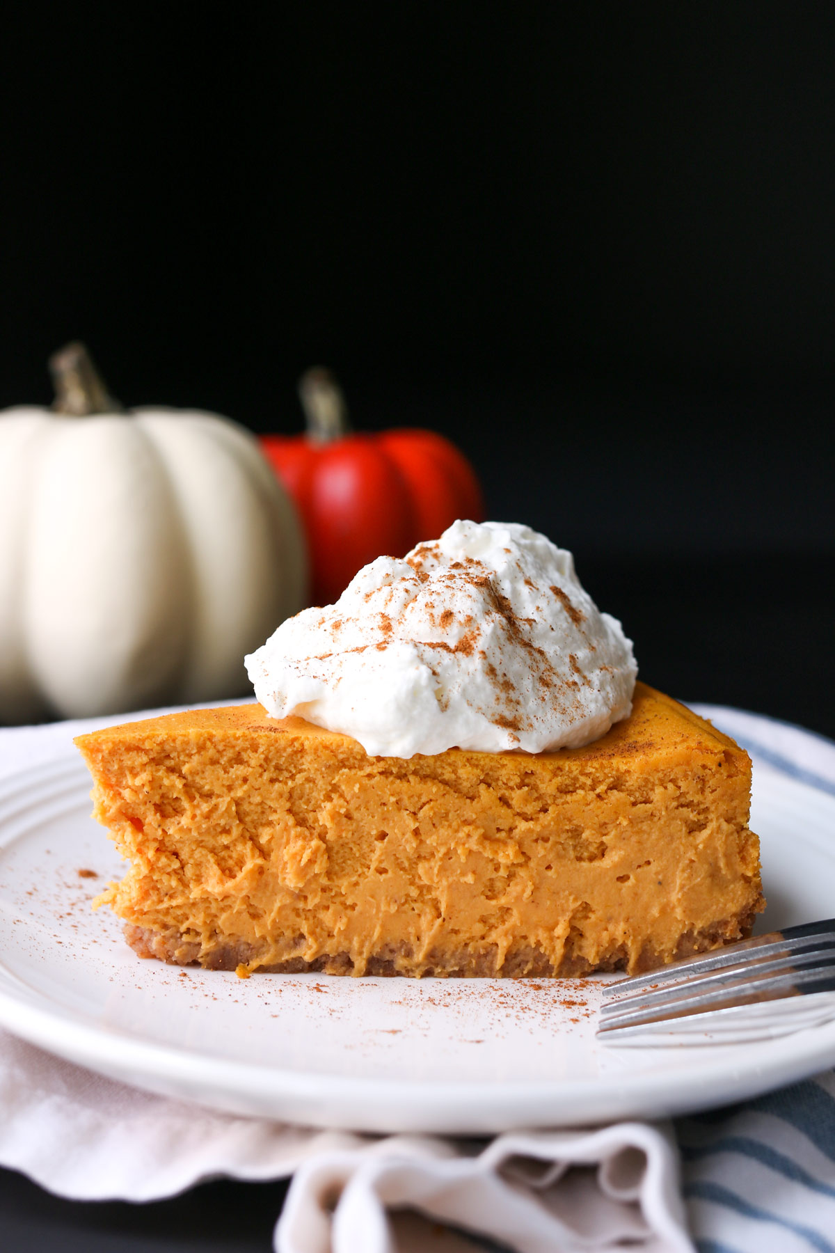 slice of cheesecake on plate with whipped cream and cinnamon, with colorful pumpkins in background.