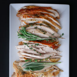 close up of sliced roast turkey breast on white rectangular platter with fresh herbs.