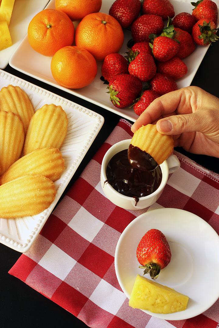 hand dipping madeleine into chocolate fondue next to plates of dippers
