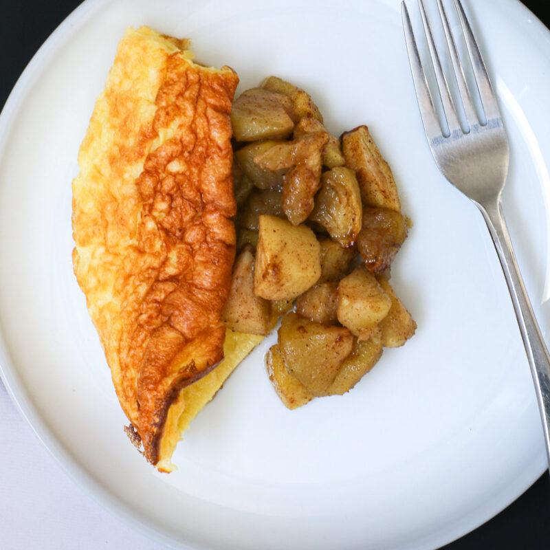 wedge of puffed pancake on plate topped with apples, a fork is nearby.